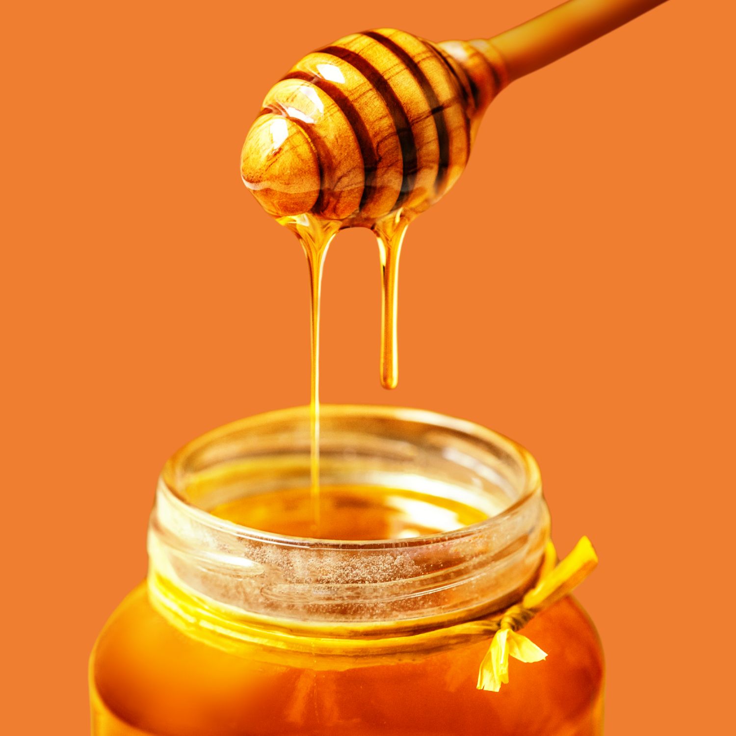 Honey drips from a wooden dipper into a glass jar filled with golden honey, set against a warm orange background.